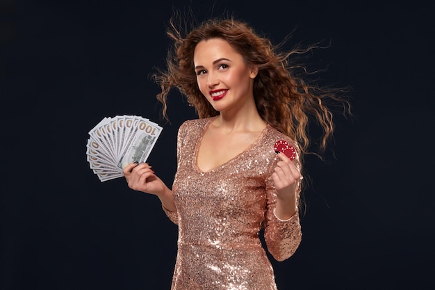 Bezpłatne zdjęcie piękna młoda szczęśliwa szczęśliwa brązowowłosa kobieta w złotej sukience koktajlowej pokazująca fan pieniędzy w jednej ręce i żetony w drugiej ręce, fan 100 dolarowych banknotów, czarne tło. hazard, kasyno,