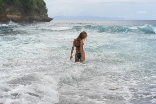 Piękna młoda szczupła kobieta z długimi blond włosami w stroju kąpielowym na plaży w pobliżu oceanu. Odpoczynek na plaży. Tropikalne wakacje. Kobieta wchodzi do wody, żeby popływać.
