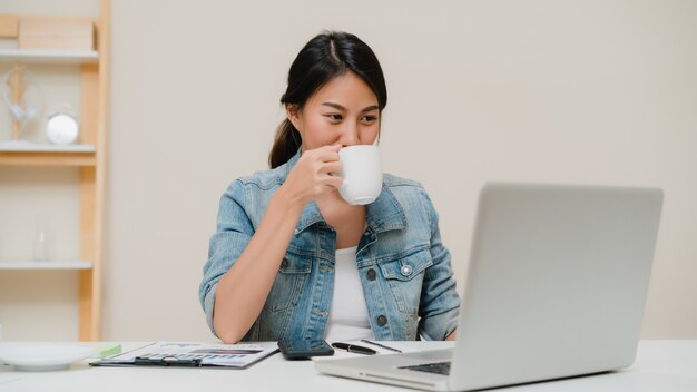 Piękna młoda uśmiechnięta azjatykcia kobieta pracuje na laptopie i pije kawę w żywym pokoju w domu.