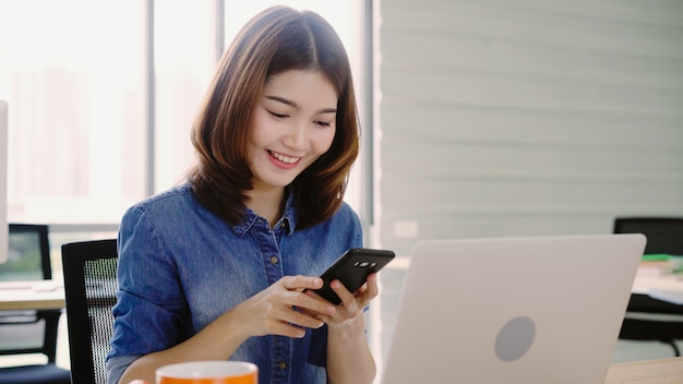 Piękna młoda uśmiechnięta azjatykcia kobieta pracuje na laptopie podczas gdy cieszący się używać smartphone przy biurem.