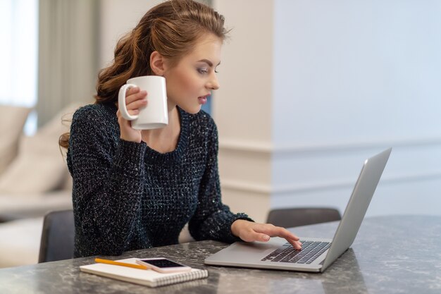 Piękna młoda uśmiechnięta kobieta pracuje na laptopie i pije kawę siedząc w domu