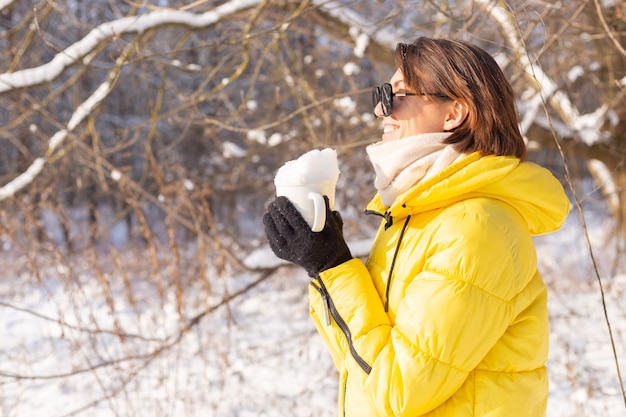 Bezpłatne zdjęcie piękna młoda wesoła kobieta w lesie zimą śnieżny krajobraz w okularach przeciwsłonecznych z kubkiem wypełnionym śniegiem, zabawy