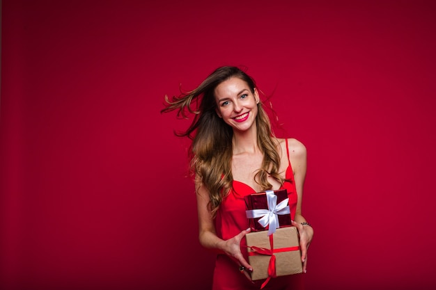 Piękna modelka w czerwonej sukience trzyma wiele małych pudełek z prezentami i raduje się