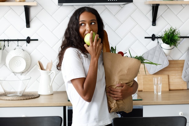 Piękna oliwkowa kobieta trzyma pakiet pełen jedzenia i je jabłko w nowoczesnej białej kuchni