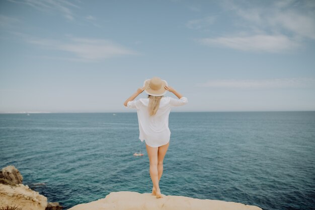 Piękna opalona kobieta w białej koszuli i słomkowym kapeluszu patrząc na ocean