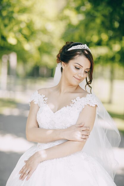 piękna panna młoda w białej sukni i koronie na głowie w parku i trzymając bukiet