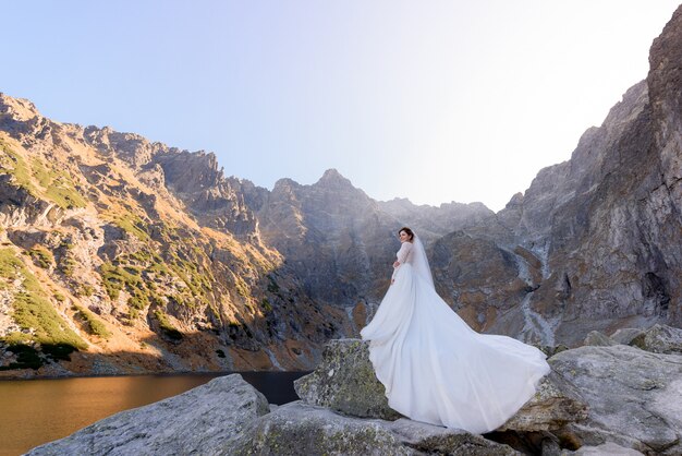 Piękna panna młoda w luksusowej sukni stoi na kamieniu w pobliżu góralskiego jeziora w ciepły słoneczny dzień