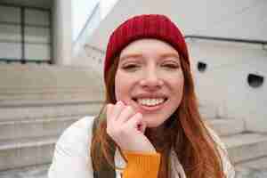 Bezpłatne zdjęcie piękna ruda studentka w czerwonym kapeluszu uśmiecha się szczerze, wygląda na szczęśliwą i zrelaksowaną, siedząc na schodach