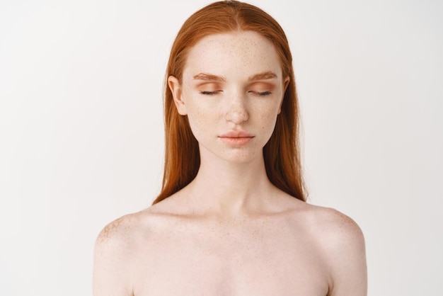 Bezpłatne zdjęcie piękna rudowłosa kobieta o bladej skórze i piegach, stojąca bez makijażu, czysta, gładka twarz, zamknięte oczy i spokojny wyraz twarzy na białym tle
