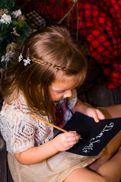 Piękna śliczna mała dziewczynka pisze list do Świętego Mikołaja w pobliżu świątecznych dekoracji na drewnianej podłodze