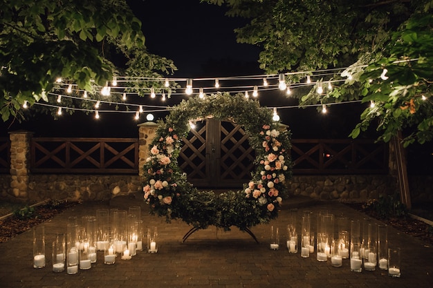 Piękna strefa fotograficzna z dużym wieńcem ozdobionym zielenią i różami na środku, świecami po bokach i girlandą zawieszoną między drzewami