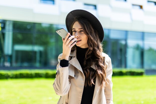 Piękna studentka rozmawia z systemem głośnomówiącym smartfona, trzymając folder na kampusie uniwersytetu.
