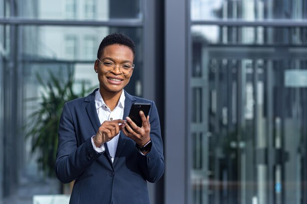 Piękna studentka trzymająca telefon, uśmiechnięta i radująca się, afroamerykanka w biznesowym stroju w pobliżu nowoczesnego biurowca