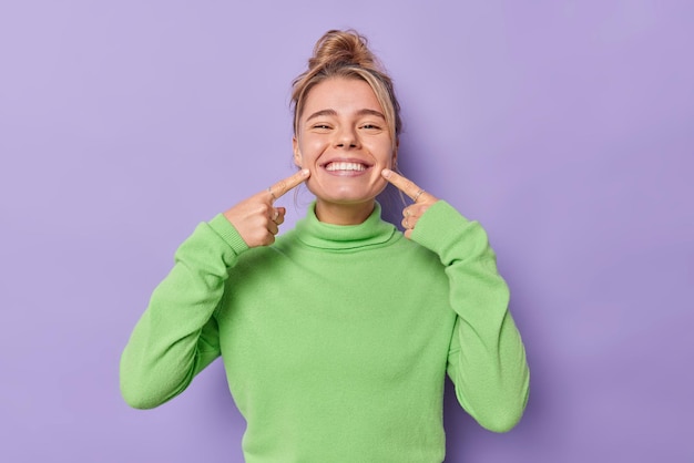Bezpłatne zdjęcie piękna szczęśliwa jasnowłosa europejka wskazuje palcem wskazującym na usta wymusza wesoły uśmiech pokazuje idealnie białe zęby nosi zielony sweterek będąc w dobrym nastroju na fioletowym tle.