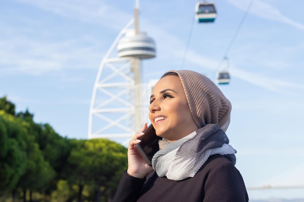 Piękna uśmiechnięta Arabska kobieta z telefonem komórkowym. Kobieta z zakrytą głową i makijaż rozmawia przez telefon komórkowy, trzymając jednorazową filiżankę kawy. Międzynarodowa, piękna koncepcja mediów społecznościowych