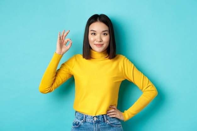 Piękna uśmiechnięta azjatycka kobieta poleca produkt, pokazując znak OK i wyglądając na zadowoloną, stojąc na niebieskim tle