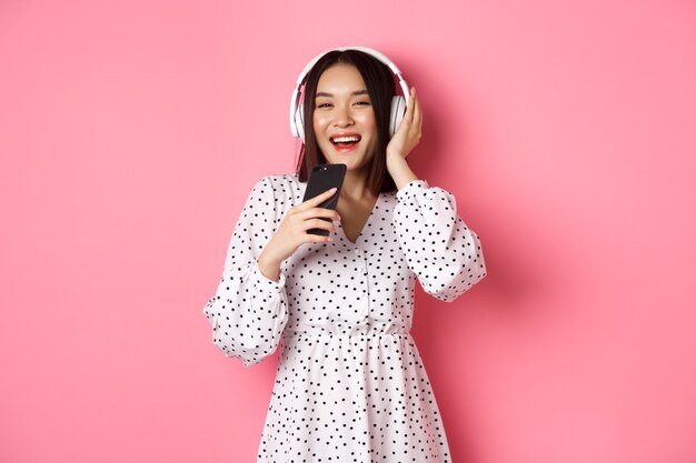 Piękna uśmiechnięta azjatycka kobieta śpiewająca piosenkę w mikrofonie smartfona, grająca w aplikację karaoke i używająca słuchawek, stojąca na różowym tle