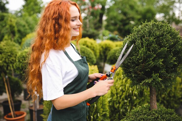 Piękna uśmiechnięta dama z rudymi, kręconymi włosami, stojąca w fartuchu i trzymająca duże nożyce ogrodowe podczas pracy na świeżym powietrzu