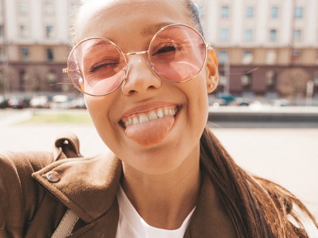 Piękna, Uśmiechnięta Dziewczyna W Lato Hipster Kurtka I Dżinsy. Model Biorąc Selfie Na Smartfonie. Kobieta Robienia Zdjęć Na Ulicy. Siedząc Na ławce W Okularach Przeciwsłonecznych I Pokazując Język