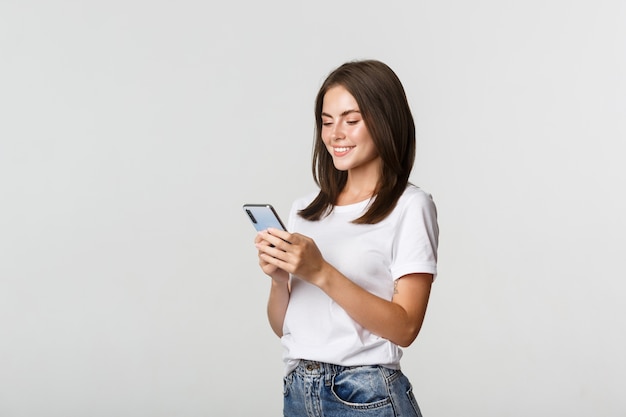 Piękna uśmiechnięta dziewczyna za pomocą telefonu komórkowego, patrząc na zadowolony smartfon.