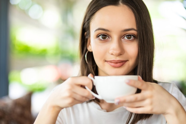 Piękna uśmiechnięta kobieta pije kawę w kawiarni. Portret dojrzałej kobiety w kawiarni, picia gorącego cappuccino i patrząc na kamery. Ładna kobieta z filiżanką kawy.
