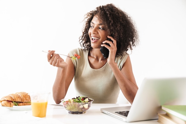 Piękna uśmiechnięta młoda afrykańska kobieta siedzi przy biurku na białym tle nad białą ścianą, jedząc lunch podczas pracy na komputerze przenośnym, rozmawiając przez telefon komórkowy