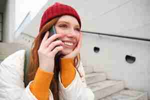 Bezpłatne zdjęcie piękna uśmiechnięta ruda modelka siedzi na ulicy i rozmawia przez telefon komórkowy za pomocą aplikacji na smartfony