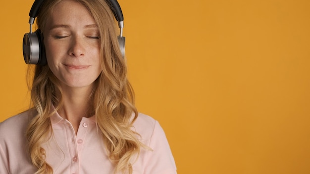 Piękna zmysłowa blond dziewczyna słucha nowej piosenki w słuchawkach, trzymając zamknięte oczy na białym tle na żółtym tle. Skopiuj miejsce na tekst lub treści reklamowe