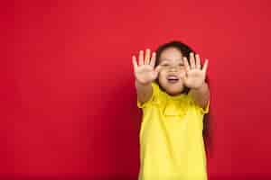 Bezpłatne zdjęcie piękne emocjonalne dziewczynki na białym tle na czerwonej przestrzeni. półdługi portret szczęśliwego dziecka pokazujący gest i skierowany ku górze. pojęcie wyrazu twarzy, ludzkie emocje, dzieciństwo.