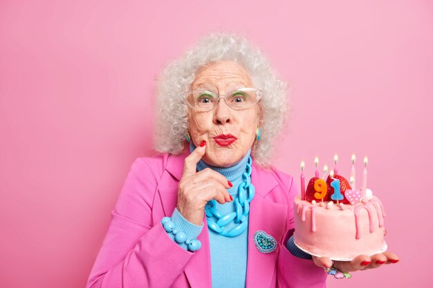 Piękne stare kręcone włosy kobieta nosi makijaż czerwona szminka ubrany w modny kostium przezroczyste okulary trzyma ciasto z płonącymi świecami świętuje jej urodziny