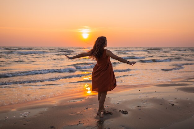 Piękne ujęcie modelu ubrana w brązową sukienkę, podziwiając zachód słońca na plaży