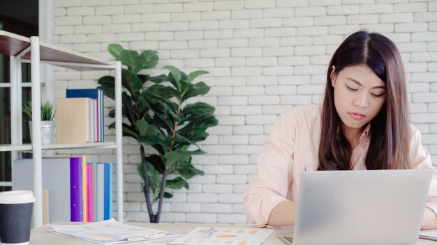 Pięknej młodej uśmiechniętej azjatykciej kobiety pracujący laptop na biurku w żywym pokoju w domu.