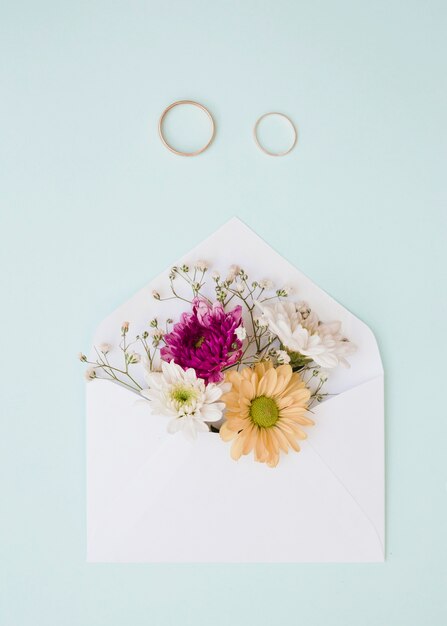 Piękni kwiaty wśrodku białej koperty z dwa obrączkami ślubnymi na błękitnym tle