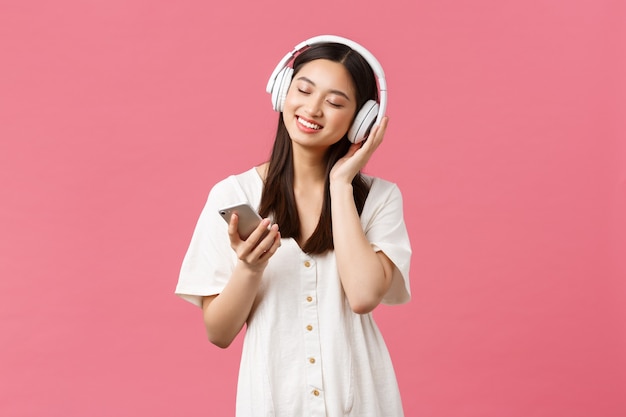 Piękno, ludzie emocje i koncepcja technologii. Beztroska zmysłowa i ładna azjatycka dziewczyna ciesząca się muzyką w bezprzewodowych słuchawkach, zamykająca oczy i uśmiechająca się słuchając ulubionej piosenki, trzymając smartfon