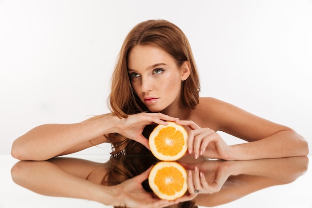 Bezpłatne zdjęcie piękno portret skoncentrowana imbirowa kobieta z długie włosy opiera na lustro stole podczas gdy trzymający pomarańczowego i patrzejący daleko od