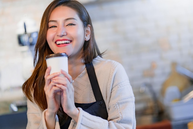 Piękny, atrakcyjny azjatycki właściciel sklepu z kawiarnią, uśmiechnięty ze szczęścia i radosny z filiżanką w fartuchu kawiarnianym tle