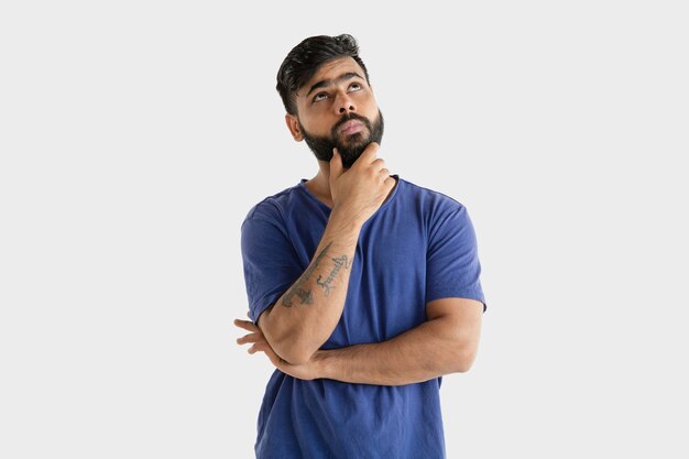 Piękny męski portret w połowie długości na białym tle na tle białego studia. Młody emocjonalny człowiek hinduski w niebieskiej koszuli. Wyraz twarzy, ludzkie emocje, koncepcja reklamy. Myślenie lub wybieranie.
