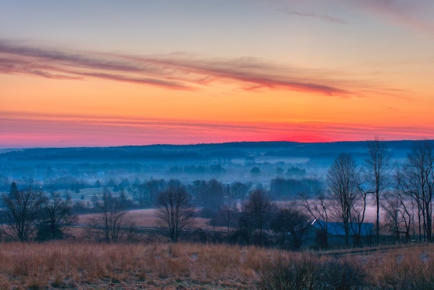 Bezpłatne zdjęcie piękny strzał niesamowitych czerwonych i pomarańczowych chmur nad dużymi mglistymi polami i lasem o świcie