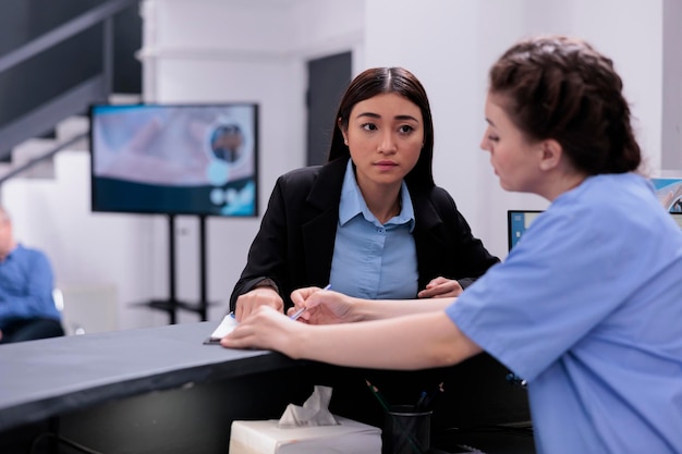 Bezpłatne zdjęcie pielęgniarka pokazująca raport medyczny recepcjonistce omawiająca ekspertyzę podczas wizyty kontrolnej w poczekalni szpitala. pracownik pomagający asystentowi w konsultacjach i wizytach kontrolnych.