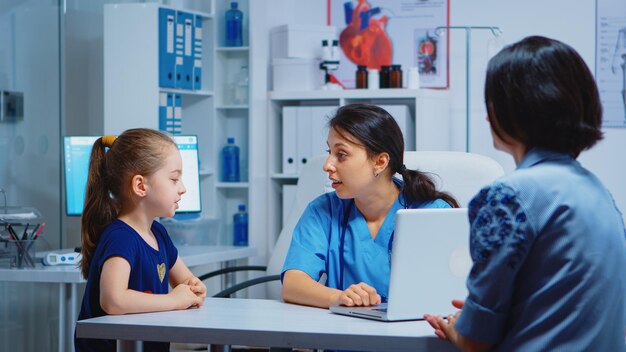 Pielęgniarka Rozmawia Z Dzieckiem I Pisze Objawy Dziewczyny Na Laptopie. Lekarz Specjalista Medycyny Udzielający świadczeń Zdrowotnych Konsultacja Badanie Diagnostyczne Leczenie W Gabinecie Szpitalnym