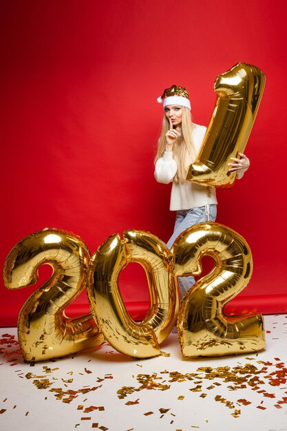 Pień fotografia blond dziewczyna w santa hat i sweter, kradnąc numer jeden nadmuchiwanej cyfry i uciekając przed cyframi Nowego Roku. Czerwone tło.