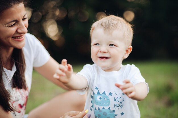 Pień fotografia pięknego chłopca w t-shirt, co pierwsze kroki w parku z matką obok. Bokeh. Niewyraźne tło. Mały chłopiec wyciągając ręce podczas chodzenia.