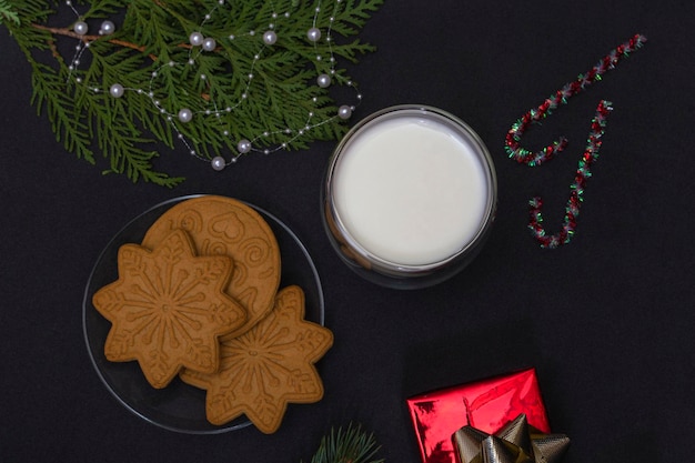 Piernik i mleko dla mikołaja. świąteczna kompozycja z piernikowymi ciasteczkami i mlekiem na czarnym tle z gałązką jodły i prezentem, widok z góry