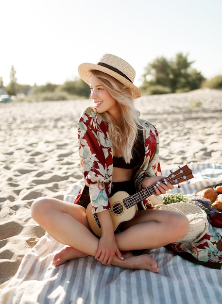Piknik na wsi. Romantyczna blondynka w słomkowym kapeluszu siedzi na okładce na plaży w delikatnych kolorach zachodu słońca i gra na gitarze ukulele. Świeże owoce, rogaliki i brzoskwinia na talerzu.