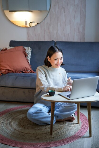 Pionowe ujęcie azjatyckiej dziewczyny siedzącej na podłodze w domu, pracującej na laptopie, studiującej w przytulnym miejscu za pomocą com