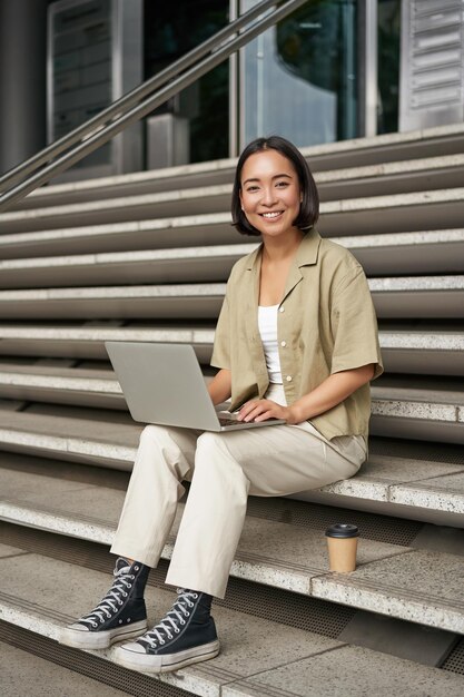 Pionowe ujęcie azjatyckiej dziewczyny siedzącej z laptopem pije kawę na schodach uniwersyteckich młoda kobieta studentka