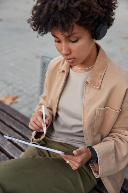 Pionowe ujęcie kobiecej freelancerki współdziałającej z ekranem tabletu używa rysika używa urządzenia z ekranem dotykowym siedzi na ławce ubranej w strój uliczny pozuje na rozmytym tle słucha muzyki w słuchawkach