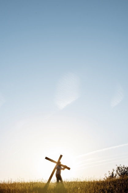 Pionowe ujęcie mężczyzny niosącego duży drewniany krzyż w trawiastym polu z błękitnym niebem