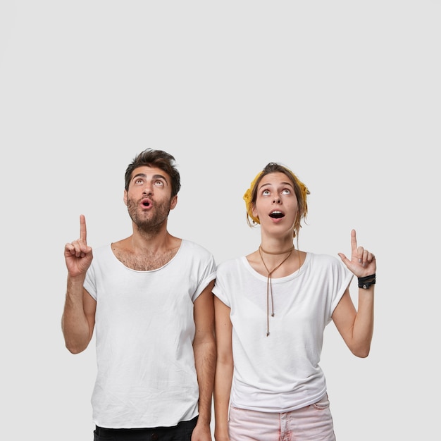 Bezpłatne zdjęcie pionowe ujęcie oszołomionej kobiety i mężczyzny wskazuje oba palce wskazujące w górę, co wskazuje na coś szokującego, opadająca szczęka, skoncentrowana na suficie