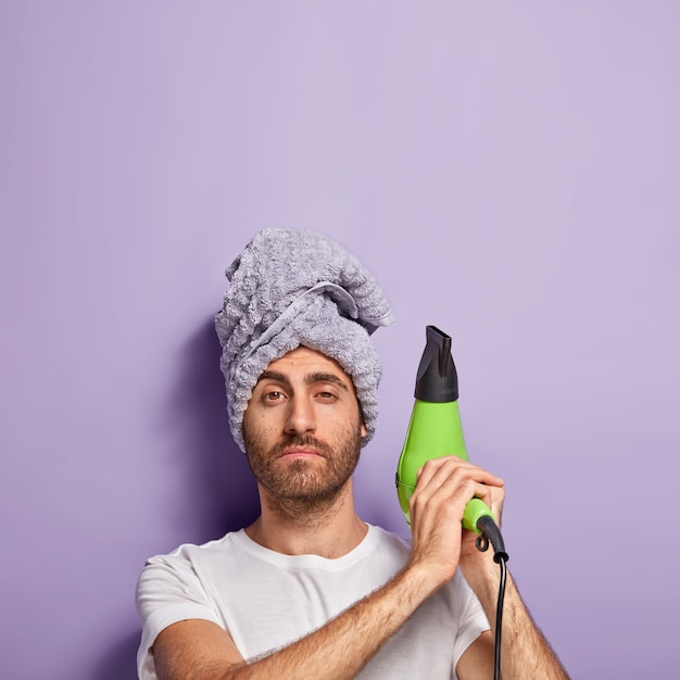Pionowe Ujęcie Pewnego Siebie Nieogolonego Mężczyzny Unoszącego Elektryczny Wiatrak, Na Głowie Nosi Ręcznik I Idzie Wysuszyć Włosy